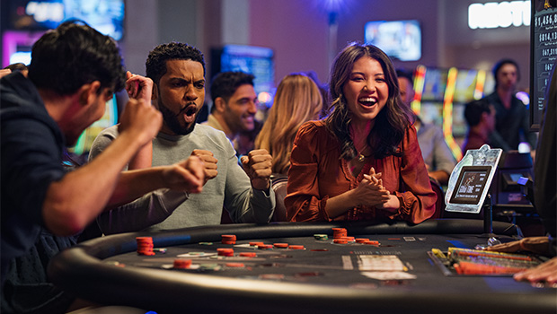A group of smiling people gathered around a poker table at a casino, with a woman in the foreground laughing as she plays.